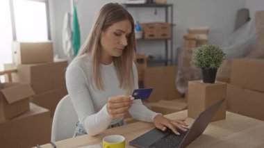 Yeni evinizde kredi kartı kullanan bir kadın masanın üzerinde bir bitki olan karton kutularla çevrimiçi alışveriş anı yakalıyor ya da yeni bir ortamda bankacılık yapıyor..