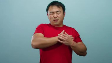 Kırmızı gömlekli adam göğsünü mavi arka planda sıkıntılı bir şekilde tutar. Gergin bir ifadeyle bir rahatsızlık veya endişe anını tasvir eder..