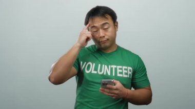 Elinde akıllı telefon tutan yeşil tişörtlü, beyaz arka planda izole edilmiş gönüllü mesajlı, düşünceli ifadeli genç adam.