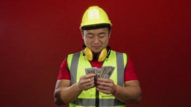 Güvenlik yeleği ve sağlam şapkalı genç Asyalı adam kırmızı arka planda kulaklıkla dolarları sayıyor..