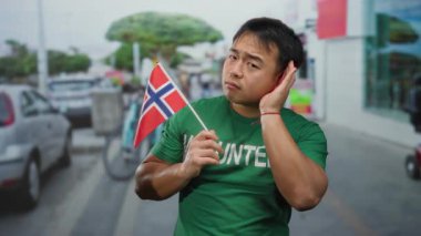 Elinde Norveç bayrağı tutan genç adam arka planı bulanık, yeşil gönüllü gömlekli, kültürel keşif ve nişanı somutlaştıran kalabalık bir şehir caddesinde jest yapıyor..