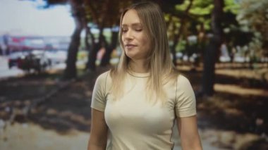Parktaki genç bir kadın, güneşli bir günde etrafı ağaçlarla çevrili, açık havada ve bulanık şehir arka planına sahip bej gömlekli, ciddi bir yüz ifadesi takınıyor..