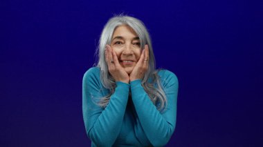 Mavi kazak giyen, gri saçlı kadın, sürpriz ve merakını dile getiren parlak mavi bir arka plana şaşırmış görünüyor..