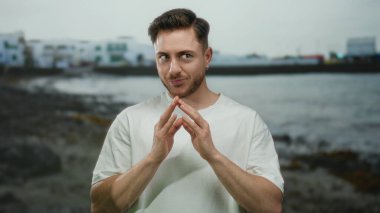 Bulutlu bir günde arka planda bulanık sahil binaları olan beyaz gömlekli genç adam deniz kenarında gülümsüyor..