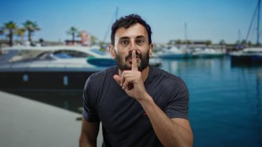 Sakallı İspanyol bir adam güneşli bir limanda sessiz kalmak için el kol hareketi yapıyor. Arka planda yatlar ve palmiye ağaçları var. Denizdeki sakin atmosferi vurguluyor..
