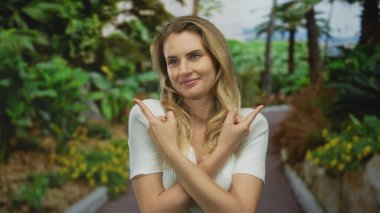 Genç sarışın kadın neşe içinde yeşil bir açık hava parkında neşeli bir ifadeyle neşeli bir yeşillik ve güneş ışığını işaret ediyor..