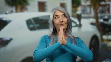 Gri saçlı ve mavi kazaklı yaşlı bir kadın işlek bir şehir caddesinde dua ediyor, araba ve şehir manzarasına karşı umut ve iyimserlik gösteriyor..