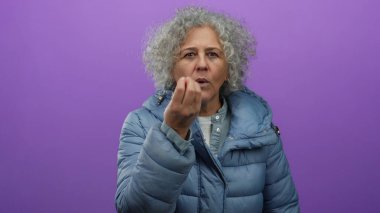 Mor arka planda mavi ceketli yaşlı bir kadın el kol hareketi yapıyor, bu da iletişimi veya ifadeyi gösteriyor, gri saçlarını ve orta yaş özelliklerini vurguluyor..