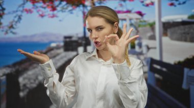 Deniz kenarındaki bir gezinti alanında, arka planda canlı çiçekler ve okyanus manzarası olan neşeli bir anda kadın jestleri yapıyor..