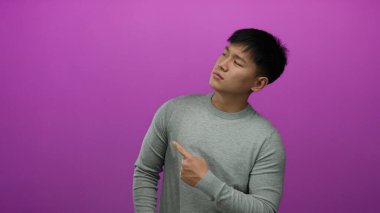 Gri kazaklı genç adam izole edilmiş pembe bir arkaplanı işaret ederek minimalist bir stüdyo ortamında merakını dile getiriyor..