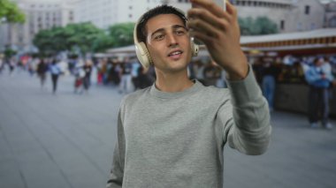 Kulaklık takan genç İspanyol adam, şehir meydanındaki işlek caddede video görüşmesi için akıllı telefon tutuyor; dijital bağlantı.