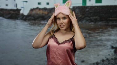 Gece elbiseli genç sarışın bir kadın arka planda deniz olan bir plajda göz maskesi takıyor, sakin bir sahil çevresi ile günlük tarzını harmanlıyor..