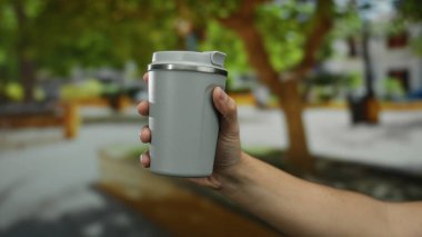 Bulanık şehir ağaçlarıyla dolu bir sokakta elinde tekrar kullanılabilir bir bardak tutan, açık hava yaşam tarzı ve sürdürülebilir seçimler sergileyen, el ve kentsel çevreye odaklanan bir adam..