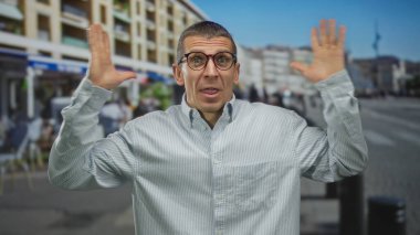 Kalabalık bir sokakta, gözlüklü ve tişörtlü, şehir binaları ve arka plandaki insanlarla çevrili bir adam şaşkınlığını ifade ediyor..