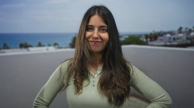Güneşli bir günde açık havada gülümseyen bir kadın sakin bir sahil atmosferi ve neşeli bir ifade sergiliyor.