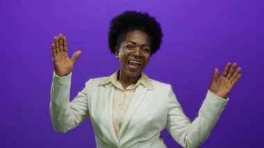 Beyaz takım elbiseli bir kadın canlı mor bir arka plana gülümsüyor, mutluluk ve profesyonellik sergiliyor, ellerini canlı bir şekilde kaldırıyor..