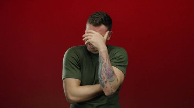 Kırmızı arka planda dövmesi olan genç bir adam yüzünü elinde tutarak hayal kırıklığını ifade ediyor..
