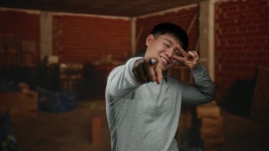 Genç adam gülümsüyor ve kapalı bir şantiyede poz veriyor. Gri bir süveterle tuğla bir duvarın önünde neşeli jestler ve neşeli bir ifade sergiliyor..