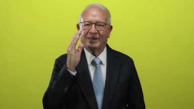 Takım elbiseli kıdemli bir adam canlı sarı bir arkaplan önünde jest yapıyor. Duygularını kendine güvenen bir iş tarzıyla aktarıyor..