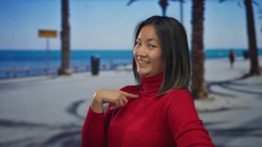 Kırmızı kazaklı genç bir kadın, arka planda palmiye ağaçları ve okyanusla deniz kenarındaki gezinti alanında gülümserken kendini işaret ediyor..