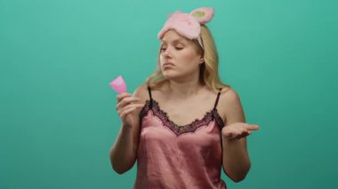 Pembe gecelikli genç sarışın kadın yeşil arka planda şüpheli bir ifadeyle regl bardağı tutuyor..