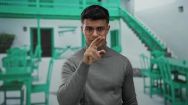 Dışarıda terasta jestler yapan genç bir adam, gözlüklü ve gri kazaklı, yeşil sandalyeli ve masalı bir şehir restoranının arka planında.