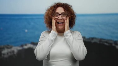 Kıvırcık esmer saçlı orta yaşlı kadın, arka planda engin mavi bir deniz ile deniz kenarında sürpriz ve neşe ifade ediyor..