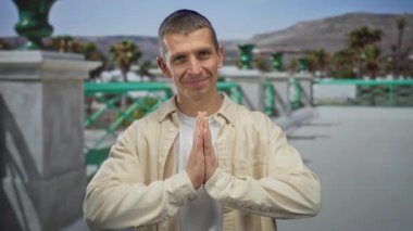 Açık havada gülümseyen beyaz bir adam. Güneşli bir sokakta, aydınlık bir gökyüzüne karşı dua eden bir jestle..