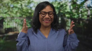 Gözlüklü ve mavi bluzlu kadın, parkta güneşli yeşil yapraklar arasında el kaldırıyor ve gülüyor; saf neşe.