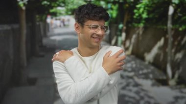 Sokakta omuzlarını kapalı tutan adam, kapalı gözler ve nazik gülümseme, beyaz süveter giyiyor ve benekli güneş ışığı altında gözlük; huzur.