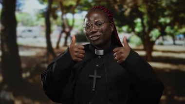 Çapraz gülümseyen ve parkın düzeninde baş parmak işareti yapan, gözlüklü ve siyah cüppeli bir kadın, doğal çevrede pozitif hava yayıyor..