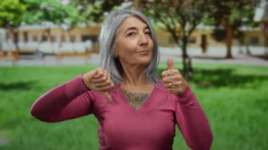 Pembe kazaklı, gri saçlı yaşlı kadın yeşil çimenler ve ağaçlarla dolu canlı bir açık hava parkında karışık duyguları baş parmaklarıyla tasvir ediyor..
