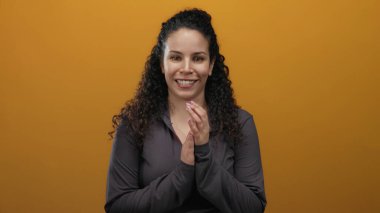 İspanyol kadın canlı turuncu bir arkaplan karşısında alkışlıyor, neşeli ve pozitif bir ifade yayıyor..