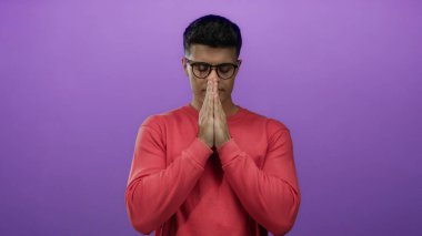 Kırmızı kazaklı genç adam mor bir arka plana karşı dua ediyor, elleri bir arada, gözleri kapalı, izole edilmiş, sessiz iç gözlem ifade ediyor..