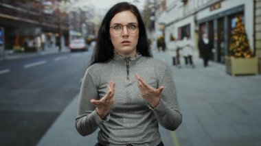 Kadın kışın şehir caddesinde, bulanık yayalar ve şehir dekorasyonlarıyla çevrili, şaşkın bir ifadeyle kendini işaret ediyor..