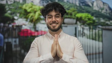 Genç, sakallı, gözlüklü, dua ederken gülümseyen, yeşil ve dağlarla çevrili genç bir İspanyol.