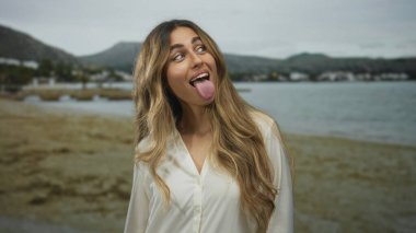 Uzun dalgalı saçları olan genç sarışın kadın kumlu bir sahil kıyısında dil sallıyor; şapşallık eğlenceli neşe oyunculuğu.