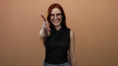 Dost canlısı bir şekilde gülümseyen bir kadın basit bir kahverengi arka plana karşı gözlüklü ve kendinden emin bir ifadeyle günlük siyah kıyafetli..