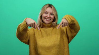 Hoş bir süveter giymiş, parmaklarıyla yeşil arka plan duvarına doğru gülümseyen bir kadın neşeli ve davetkar bir ifade yansıtıyor.