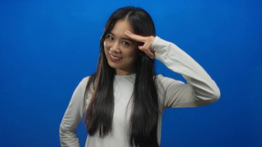 Genç Çinli bir kadın, parmakları kafasında selam veriyor mavi arka plan duvarına karşı duruyor oyuncu ve kendinden emin bir ifade sergiliyor..