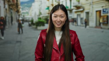 Kırmızı ceketli bir kadın arka planı bulanık, şehir yaşamını ve enerjik enerjisini gösteren hareketli bir caddede gülümsüyor..