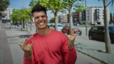 Kırmızı gömlekli genç adam şehir arka planında komik jestler yapıyor ve açık mavi gökyüzü altında modern binalar, neşeli bir güven ifadesi sergiliyor..