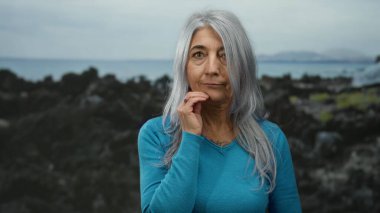 Mavi kazaklı yaşlı kadın kayalık bir sahil sahilinde dikiliyor. Düşünceli ve sakin görünüyor. Okyanus dalgaları ve bulutlu gökyüzü var..