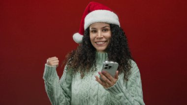 Kırmızı arka planda Noel Baba şapkası takan bir kadın, tatil keyfi ve şenlik havasında, rahat bir kazakla akıllı telefon tutarken görülüyor..