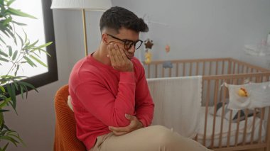 Genç adam düşünceli bir şekilde bir beşiğin yanındaki odada oturuyor, gözlük takıyor ve kırmızı bir kazak giyiyor, tefekkür ve iç gözlem hissini yansıtıyor..