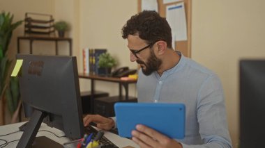 Ofiste tablet ve bilgisayar kullanan sakallı İspanyol bir adam, odaklanmış çalışma ortamı için bitkiler, kitaplar ve kırtasiye malzemeleriyle çevrili..