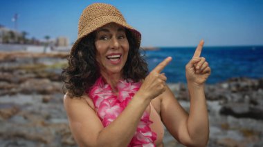 Orta yaşlı, bikinili, açık havada güneşli bir günde Hawaii lehçesi ve şapkası olan, gülümseyen ve sahili gösteren İspanyol bir kadın..