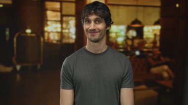 Çıplak kollu bir adam gülümsüyor ve bir otel binasında yük arabasının yanında duruyor. Gri bir tişört giyiyor..