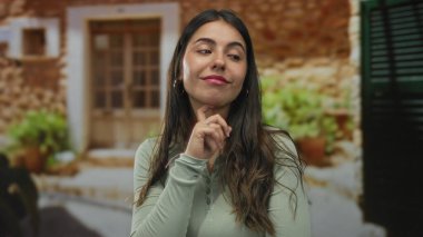 Kırsal alanda düşünceli bir şekilde gülümseyen genç İspanyol kadın. Arka planda taş duvarlar ve yeşillik var. Huzurlu ve düşünceli bir ruh hali sergiliyor.