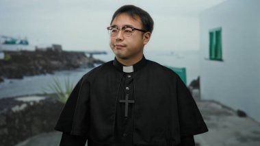 Siyah cüppeli rahip deniz kenarındaki gezinti alanında gözlüklü ve arka planda çapraz olarak görülebilecek şekilde stresli görünüyor.